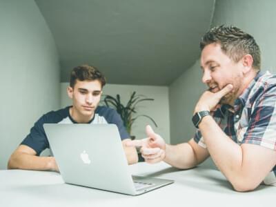 Man points at laptop screen as young man watches on